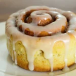 A close-up of a sourdough discard cinnamon rolls roll topped with creamy icing, placed on a white plate.