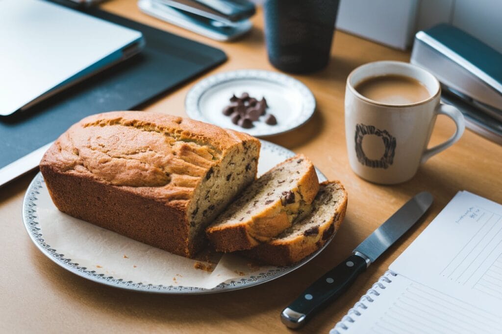 Sliced banana bread with chocolate chips on a plate next to a cup of coffee, surrounded by office supplies.